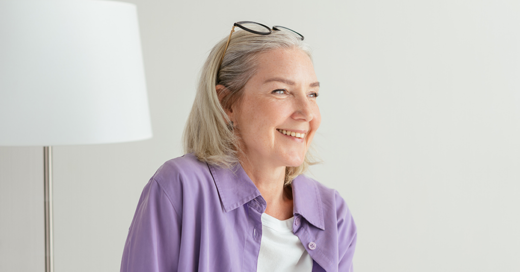 An attractive older woman with shorter hair, wearing a light purple shirt, and smiling looking off to the side